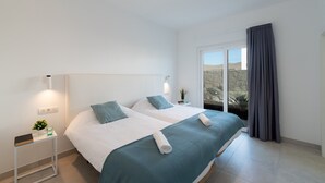 A modern bedroom with two beds, a bedside table with a plant, and a view of the outdoors through a large window. - Villa Malvasia by Villa Plus (Playa Blanca)