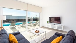 A modern living room with a pool view, a flat-screen TV, and a sofa with cushions. - Villa Mifaya by Villa Plus (Playa Blanca)