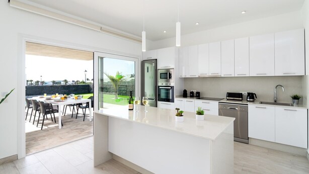 A modern kitchen with white cabinetry, a central island, and a dining area with a view of the outdoors. - Villa Mifaya by Villa Plus (Playa Blanca)