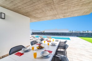 Zona pranzo all'aperto con vista piscina, tavolo apparecchiato con piatti, bicchieri e una ciotola di frutta.