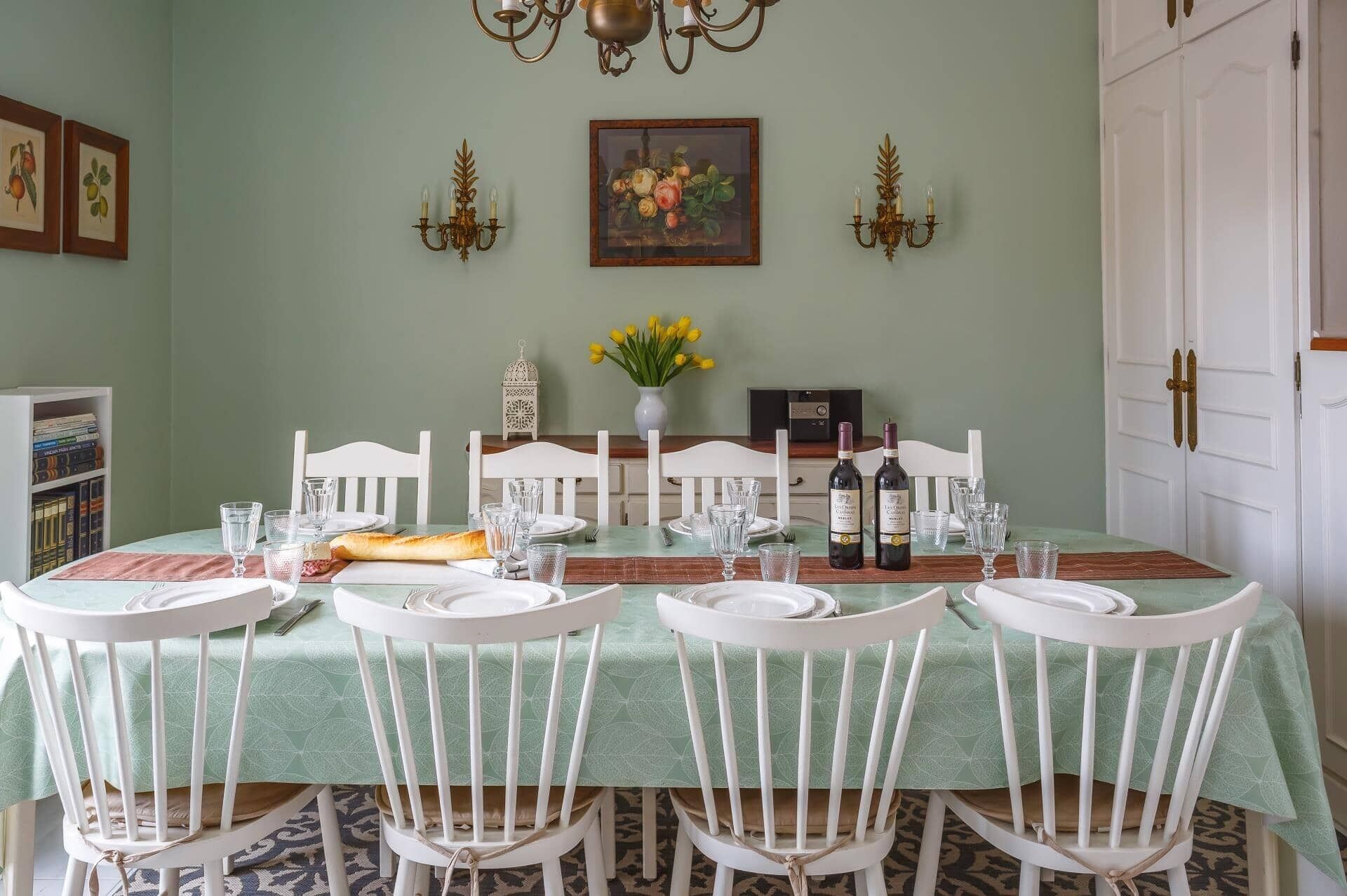 Une salle à manger avec une nappe verte, des chaises blanches et un lustre. Des tableaux encadrés ornent le mur, et un vase rempli de fleurs est posé sur la table.