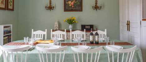 Une salle à manger avec une nappe verte, des chaises blanches et un lustre. Des tableaux encadrés ornent le mur, et un vase rempli de fleurs est posé sur la table.
