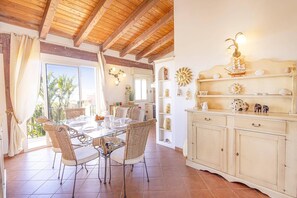 Un comedor con mesa de vidrio, sillas de mimbre, techo de madera y un mueble decorativo con estantes.