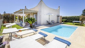 Un espace aménagé au bord de la piscine, avec des chaises longues, un coin salon abrité et un bâtiment blanc doté d’une conception architecturale originale, notamment au niveau du toit.