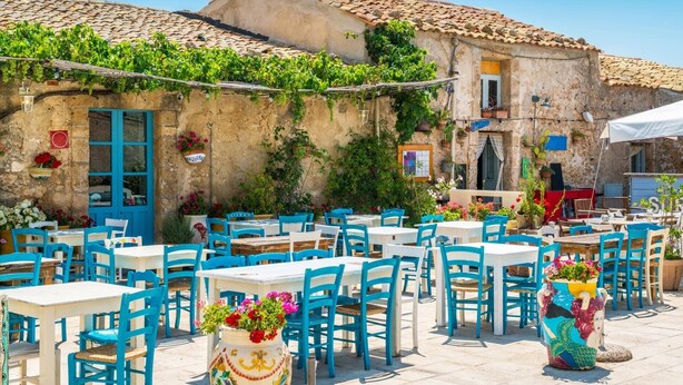 Un café en plein air avec des chaises et des tables bleues, entouré d’un bâtiment en pierre doté d’un toit en tuiles et agrémenté de verdure.