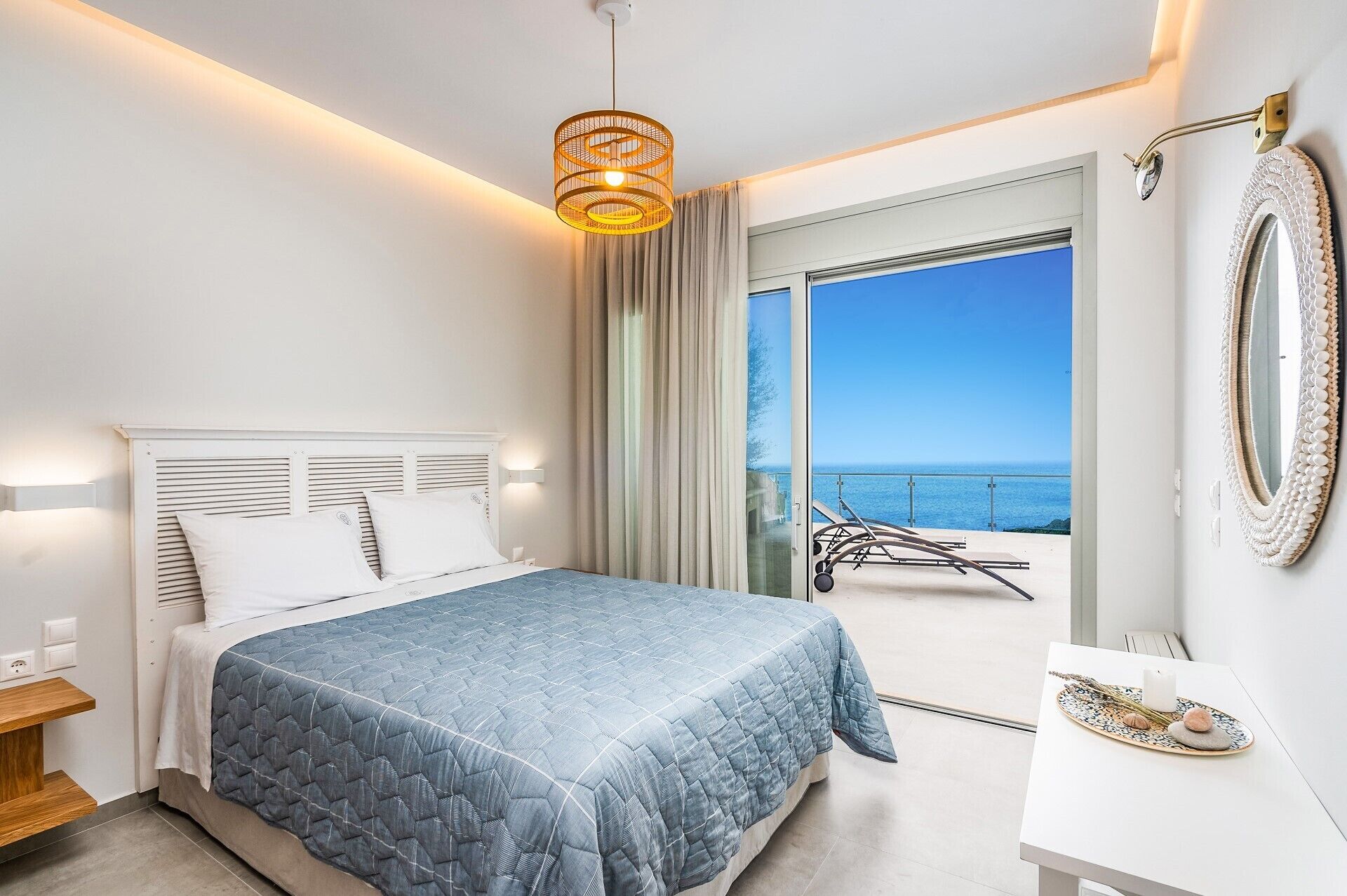 A bedroom with a bed, a bedside table, a nightstand, a large window with a view of the sea, and a balcony with a chair and table.