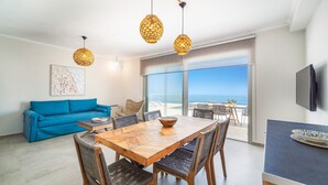 Ein modernes Wohnzimmer mit einem blauen Sofa, einem Holzespeisesaal mit Korbstühlen und einem großen Fenster mit Meerblick.