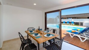 Zona pranzo con tavolo imbandito per quattro persone, vista sulla piscina e un edificio moderno sullo sfondo.