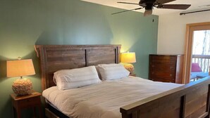 Een slaapkamer met een houten bed, twee bedlampen, een plafondventilator en uitzicht op het omliggende landschap door een raam.