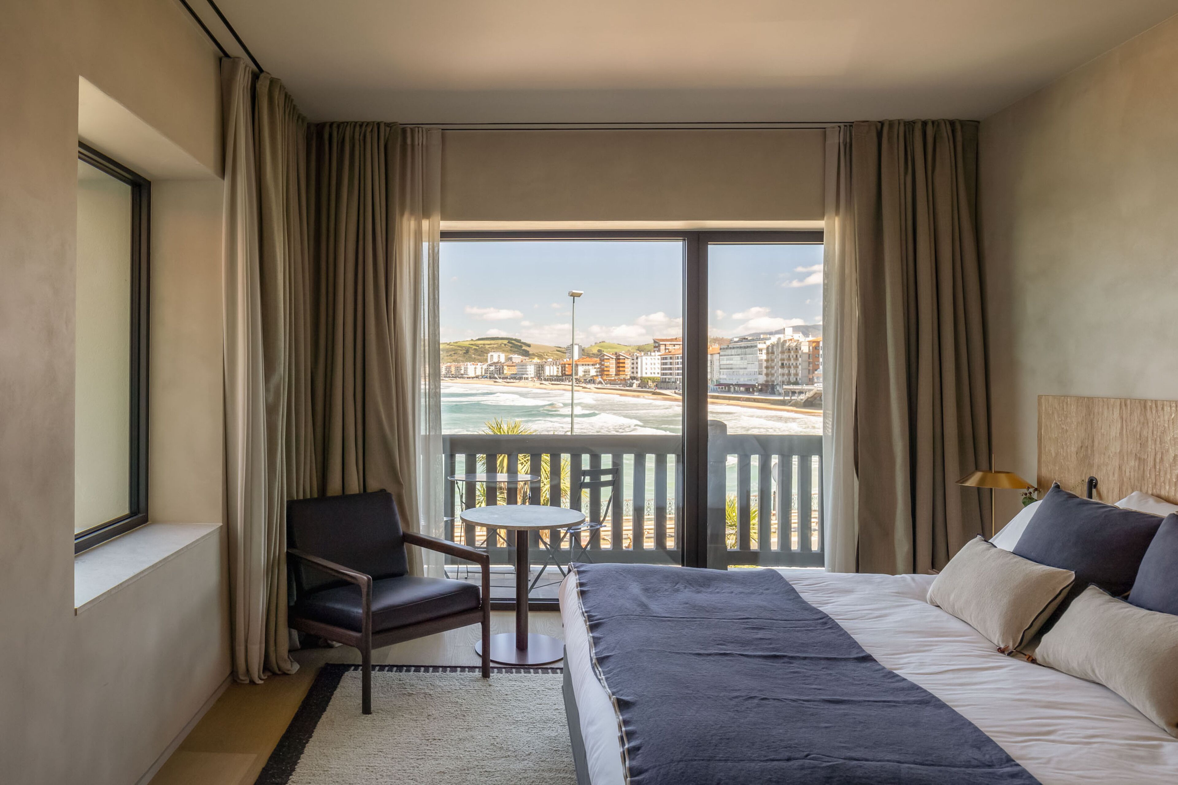 Hotelkamer met een groot bed, een stoel, een klein tafeltje en een balkon met uitzicht op het strand en gebouwen.