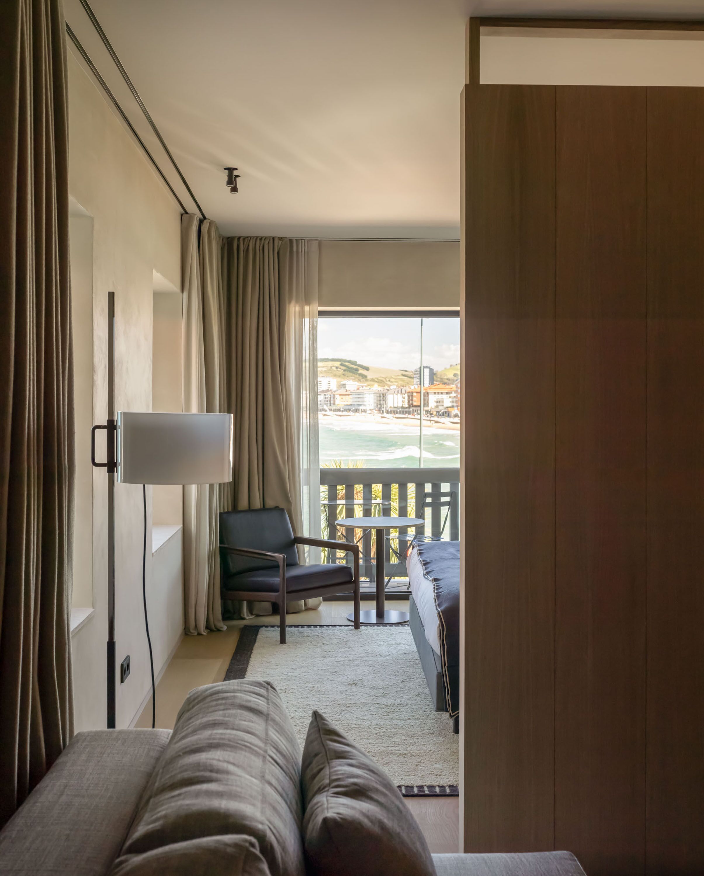 Hotelkamer met een bed, bureau, stoel en balkon met uitzicht op strand en gebouwen.
