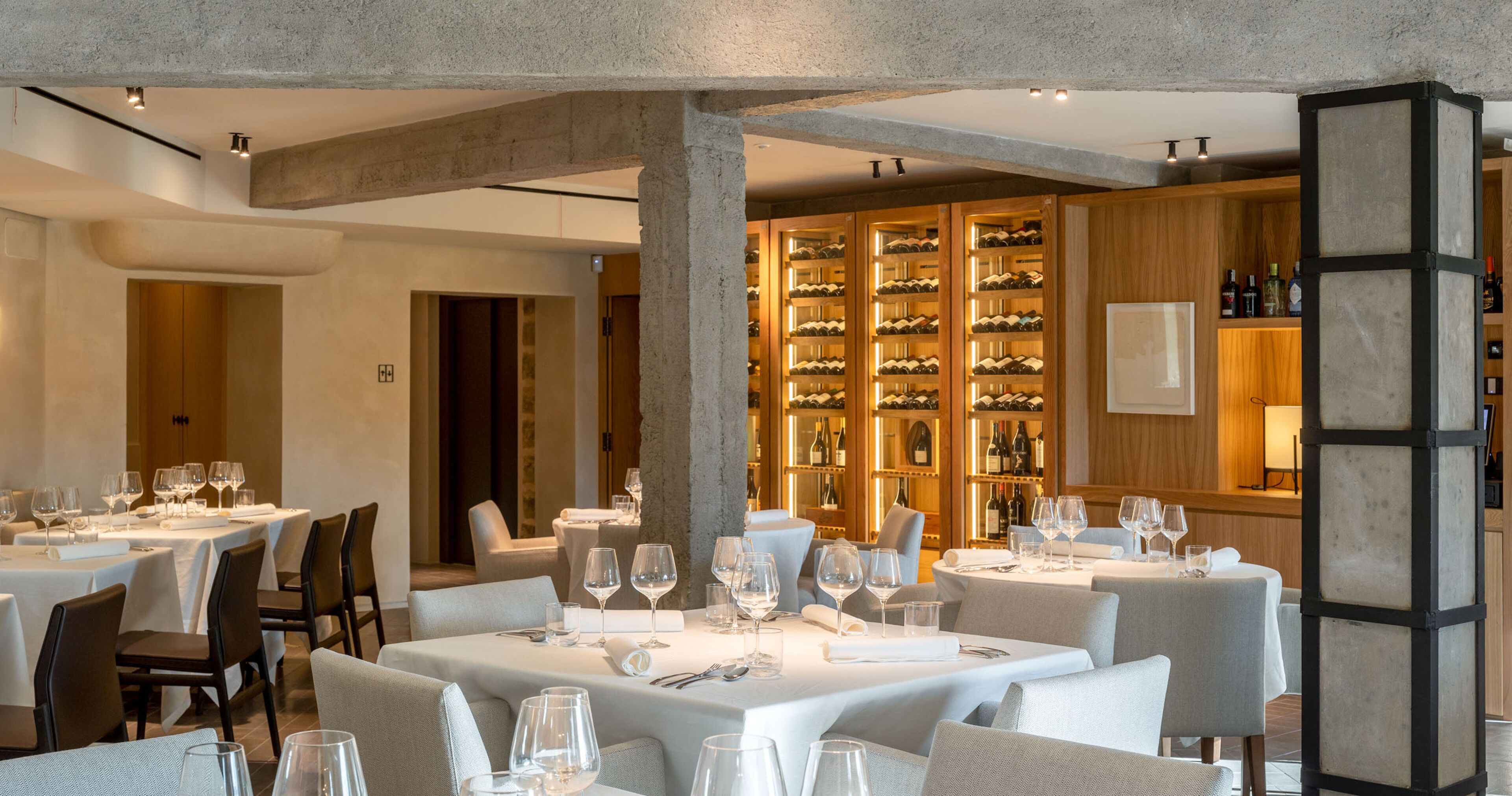 A restaurant with multiple tables set for dining, a wine cellar in the background, and a modern architectural design with exposed concrete beams.
