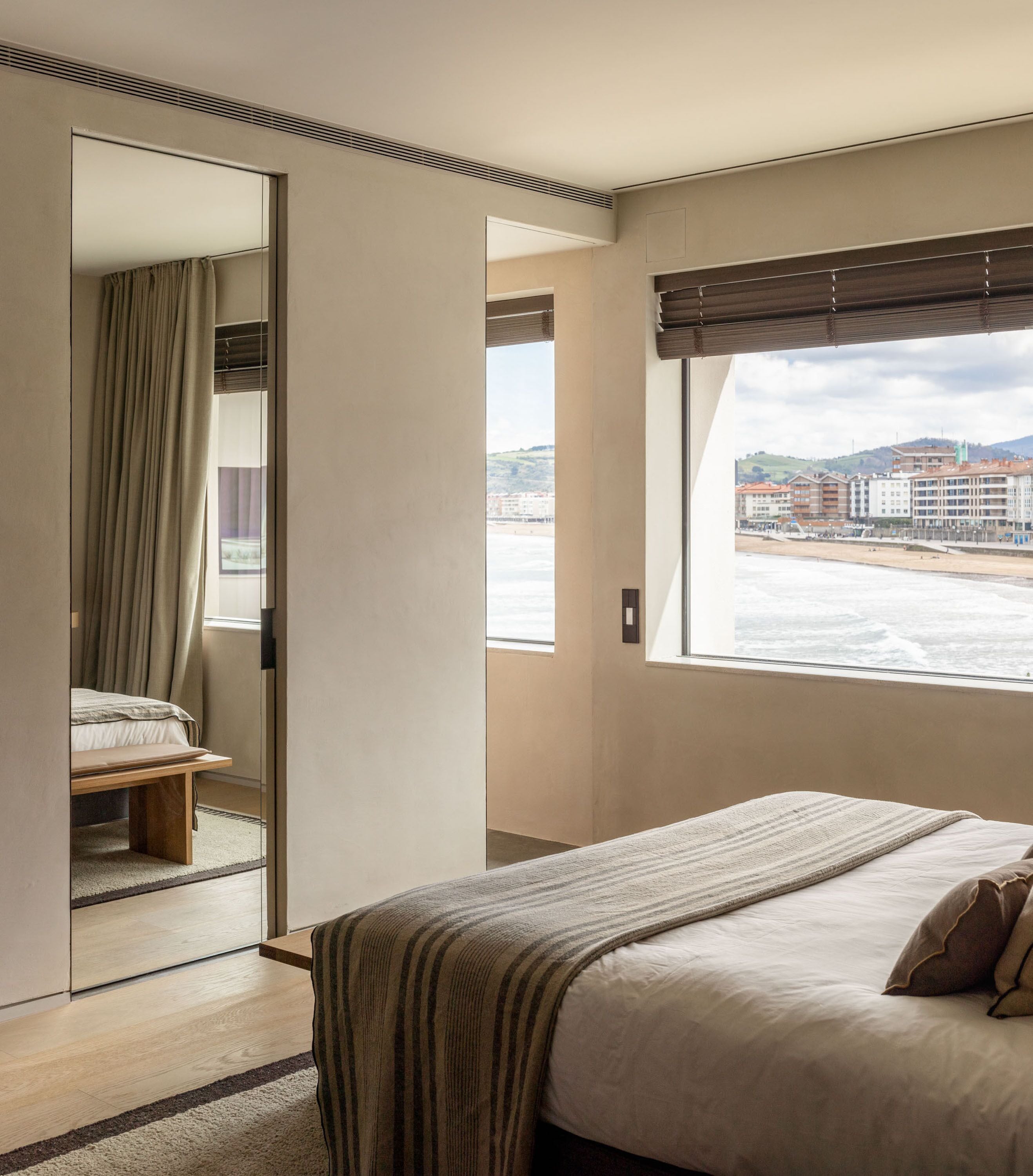 A bedroom with a bed, a bedside table, a window with a view of buildings, and a door leading to another room.