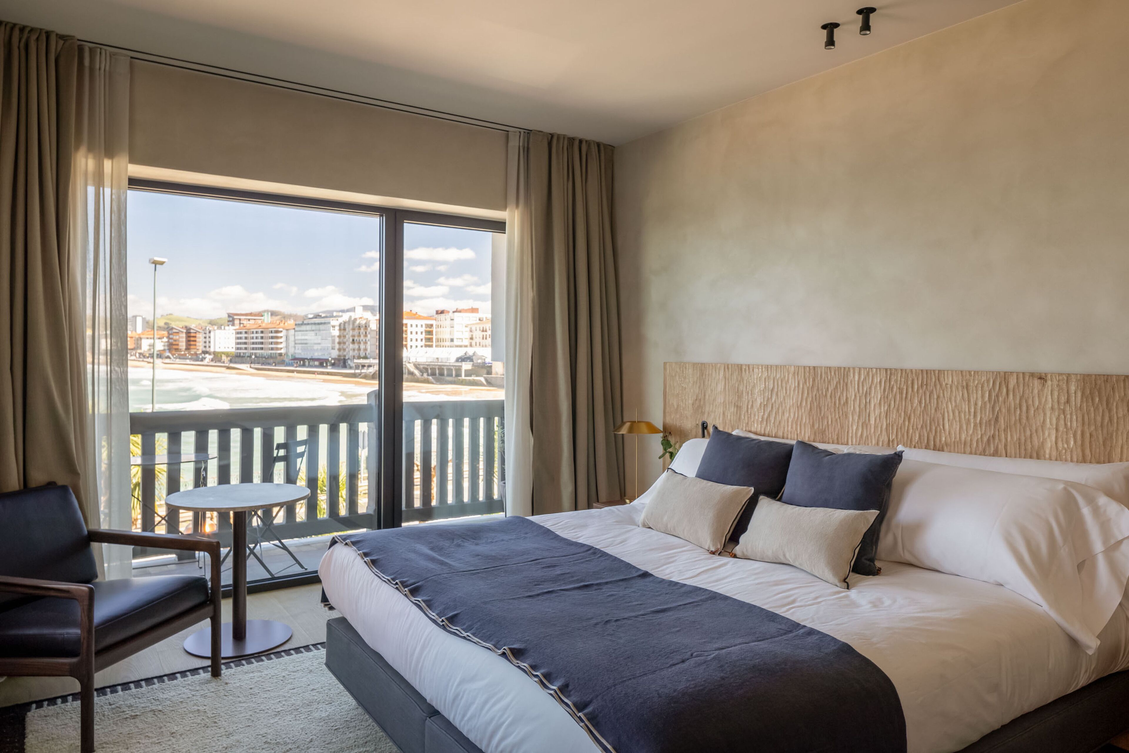 Habitación de hotel con una cama grande, una mesita, una silla y un balcón con vistas a edificios.