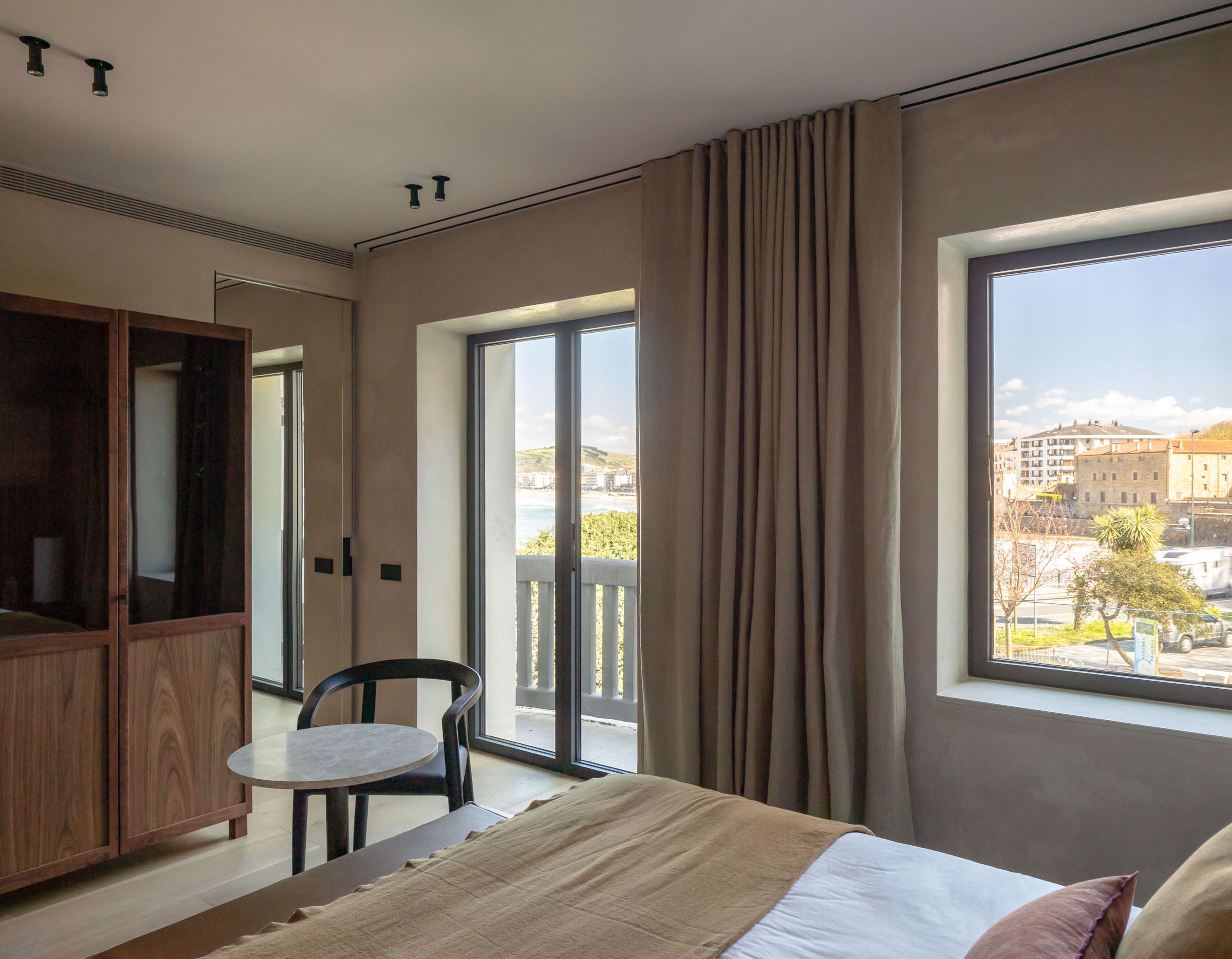 A modern hotel room with a large bed, a small table, a wardrobe, and a balcony with a view of the city.