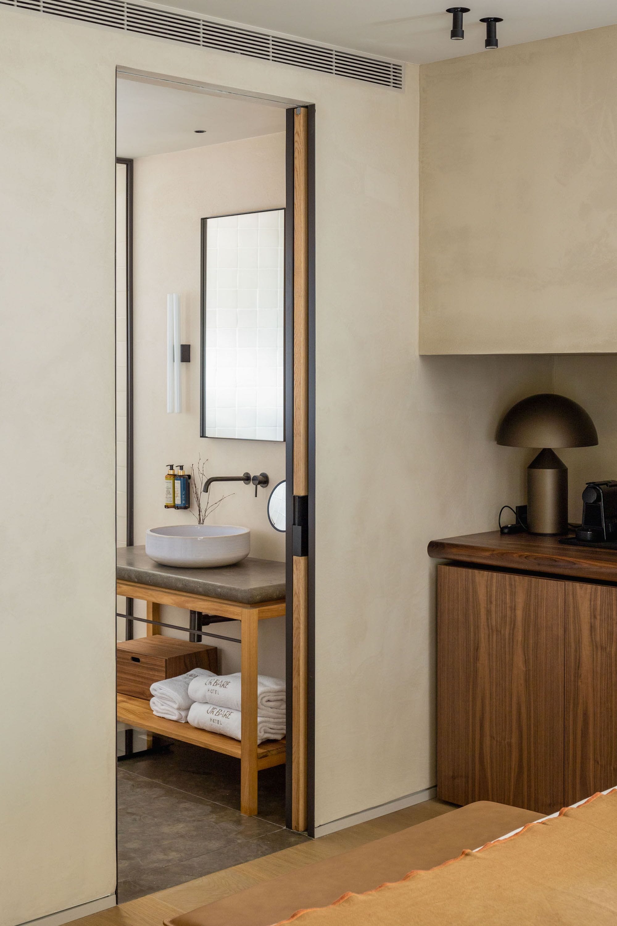 Un bany modern amb un tocador de fusta, un mirall gran i un lavabo. Hi ha un suport de tovalloles amb tovalloles doblegades i una petita estant amb productes de tocador.