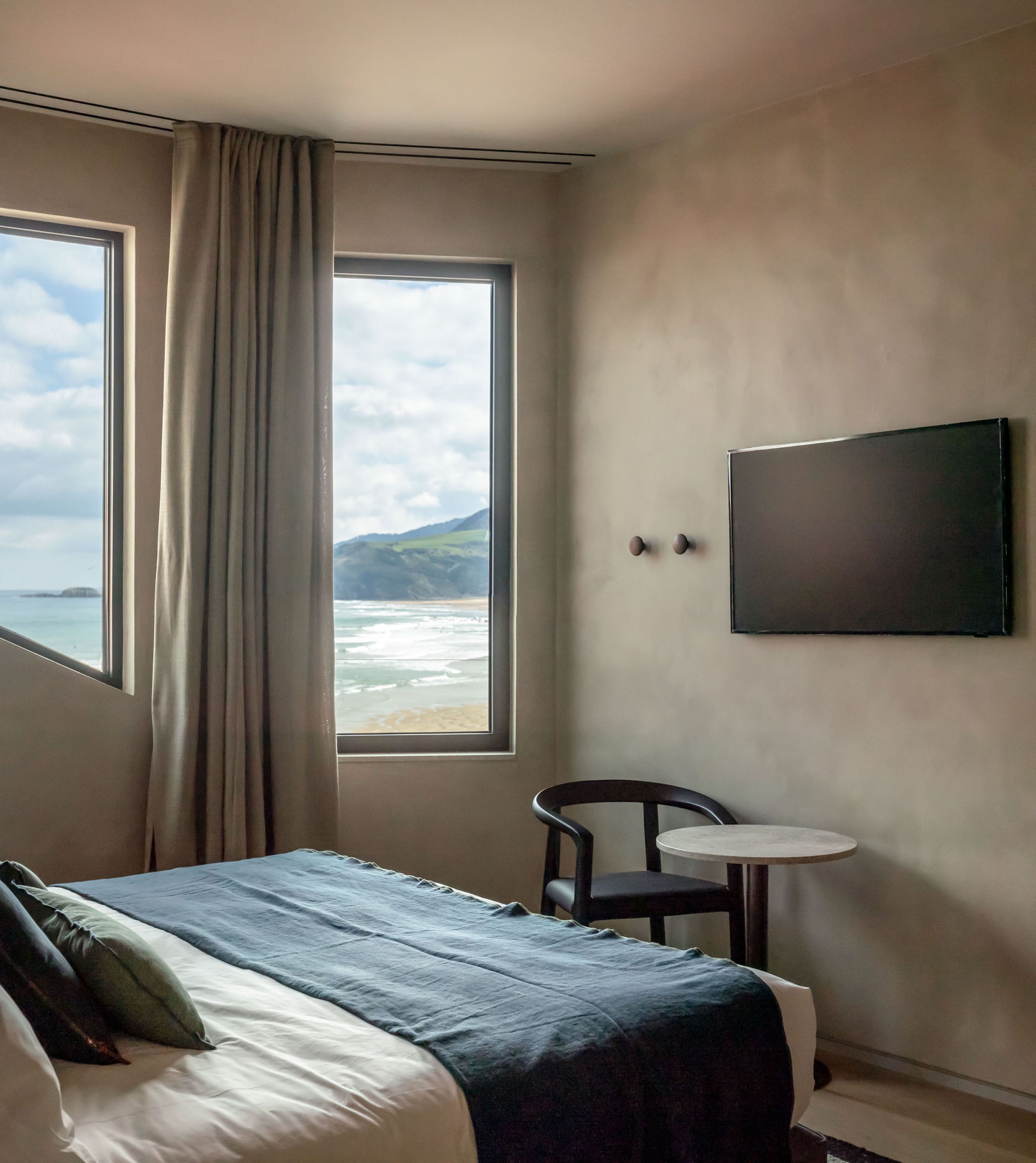 A hotel room with a bed, a chair, a small table, a TV, and a window with a view of the sea and hills.