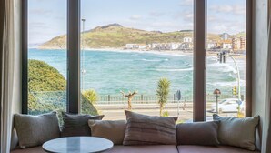 A room with a large window overlooking a coastal view, a sofa with cushions, and a small round table. - Hotel Ur Bare (Zarautz)