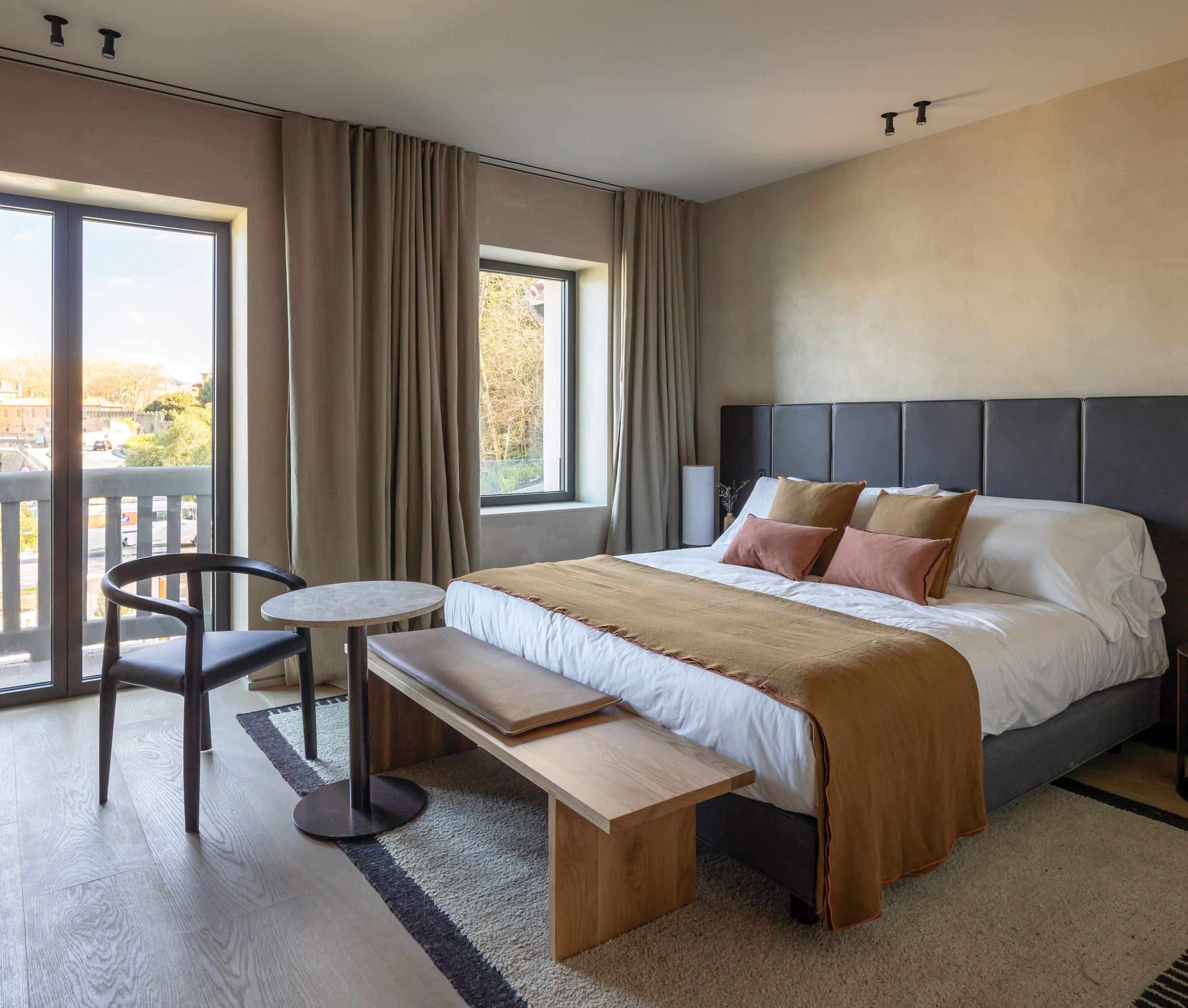 Habitación de hotel moderna con una cama grande, un banco, una mesita y vistas al exterior a través de un ventanal amplio.