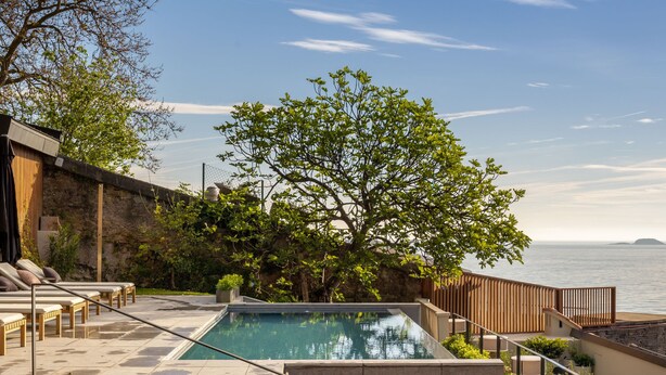 A pool area with steps leading to a lounge area, surrounded by greenery and overlooking a body of water. - Hotel Ur Bare (Zarautz)