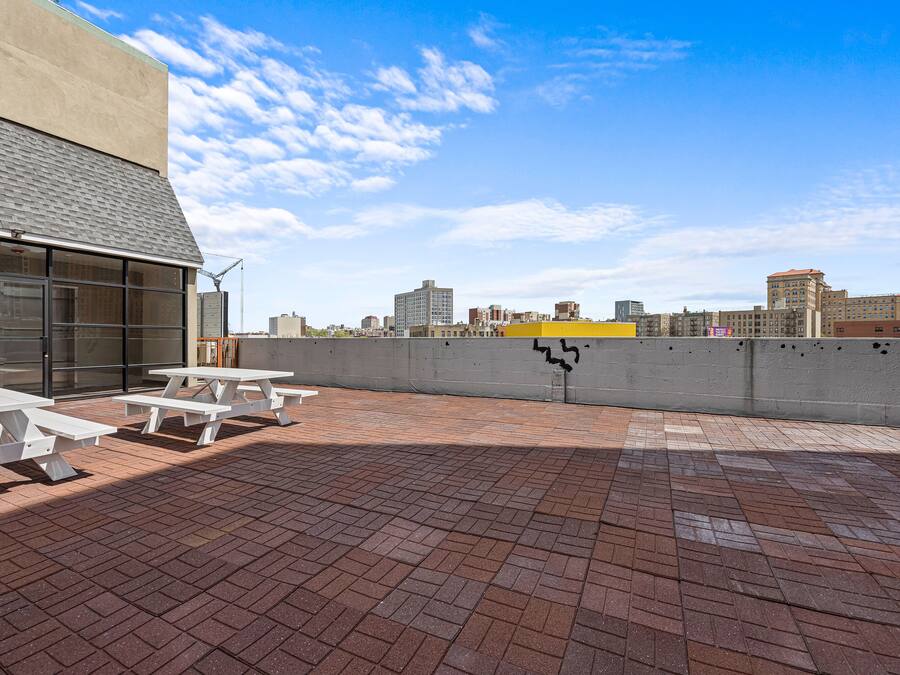 An outdoor patio with brick flooring and white picnic tables, overlooking a cityscape with buildings and a clear sky.