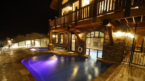 Ein großer, beleuchteter Swimmingpool mit einer Steinterrasse, umgeben von einer Holzdielen-Terrasse und Steinmauern.