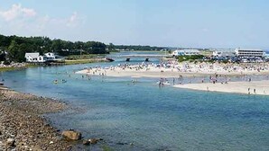 A coastal beach area with a large sandy beach, a small island with buildings, and clear blue water.