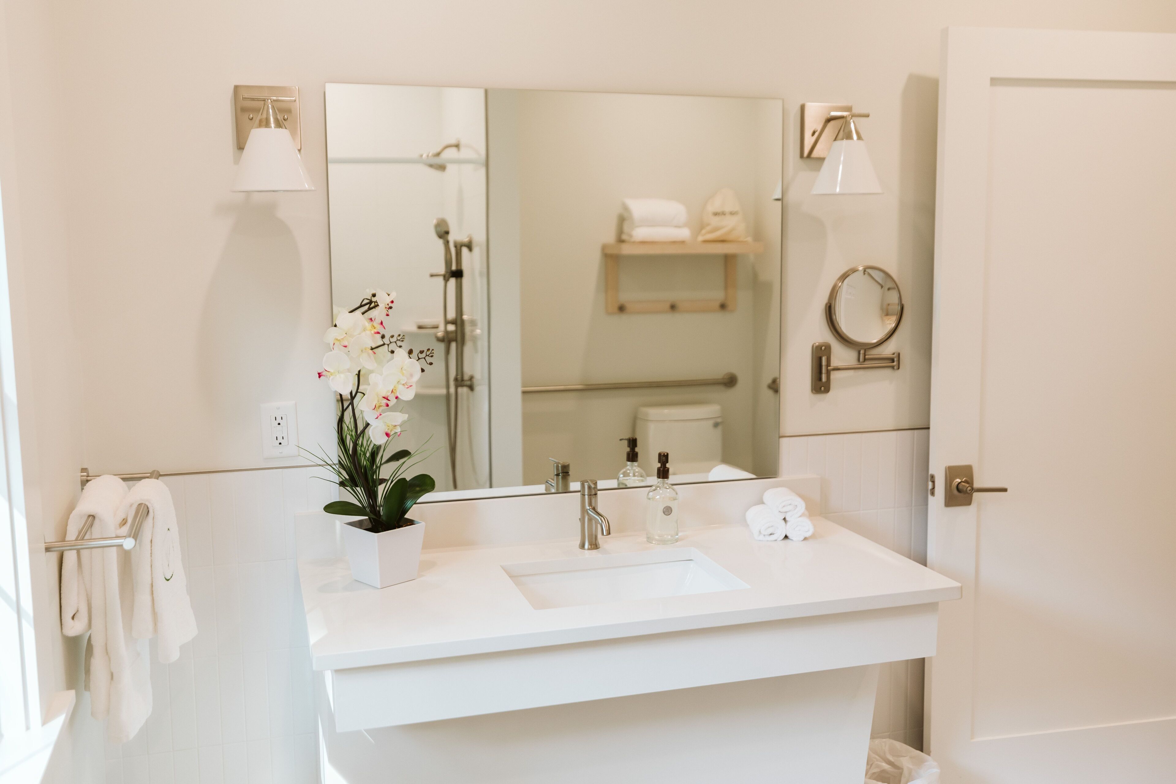 A modern bathroom with a white sink, two mirrors, a potted orchid, and a towel rack.