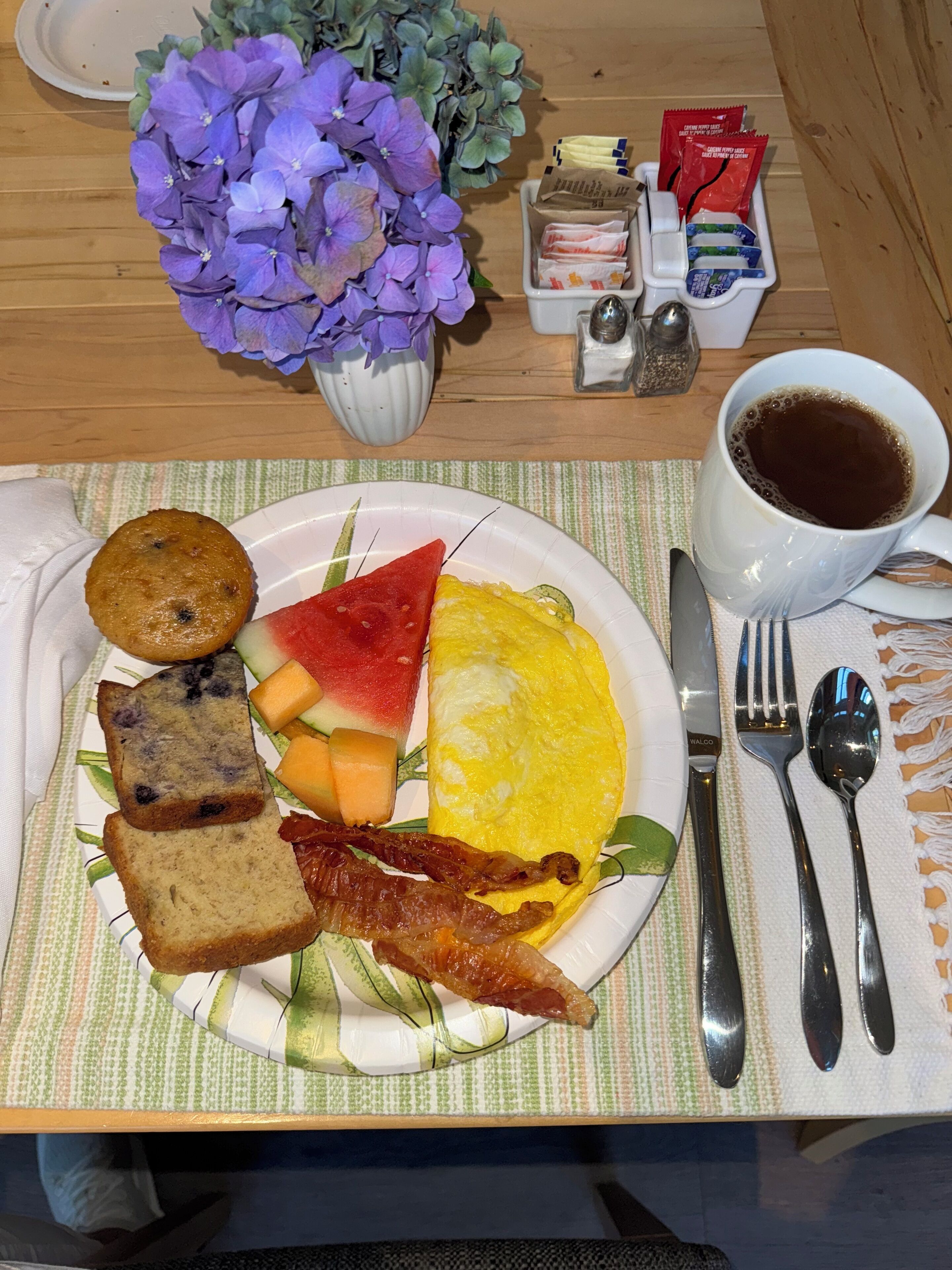 Een ontbijtbord met toast, spek, een stuk watermeloen, een plakje kaas en een gebakken ei op een tafel met een bloevas, een kop koffie en toevoegingen.