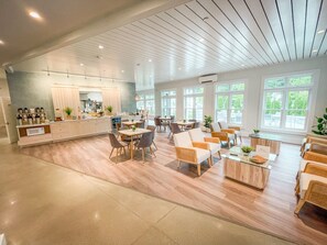Um amplo hall de hotel com área de refeições central, vários conjuntos de assentos e grandes janelas que proporcionam luz natural.