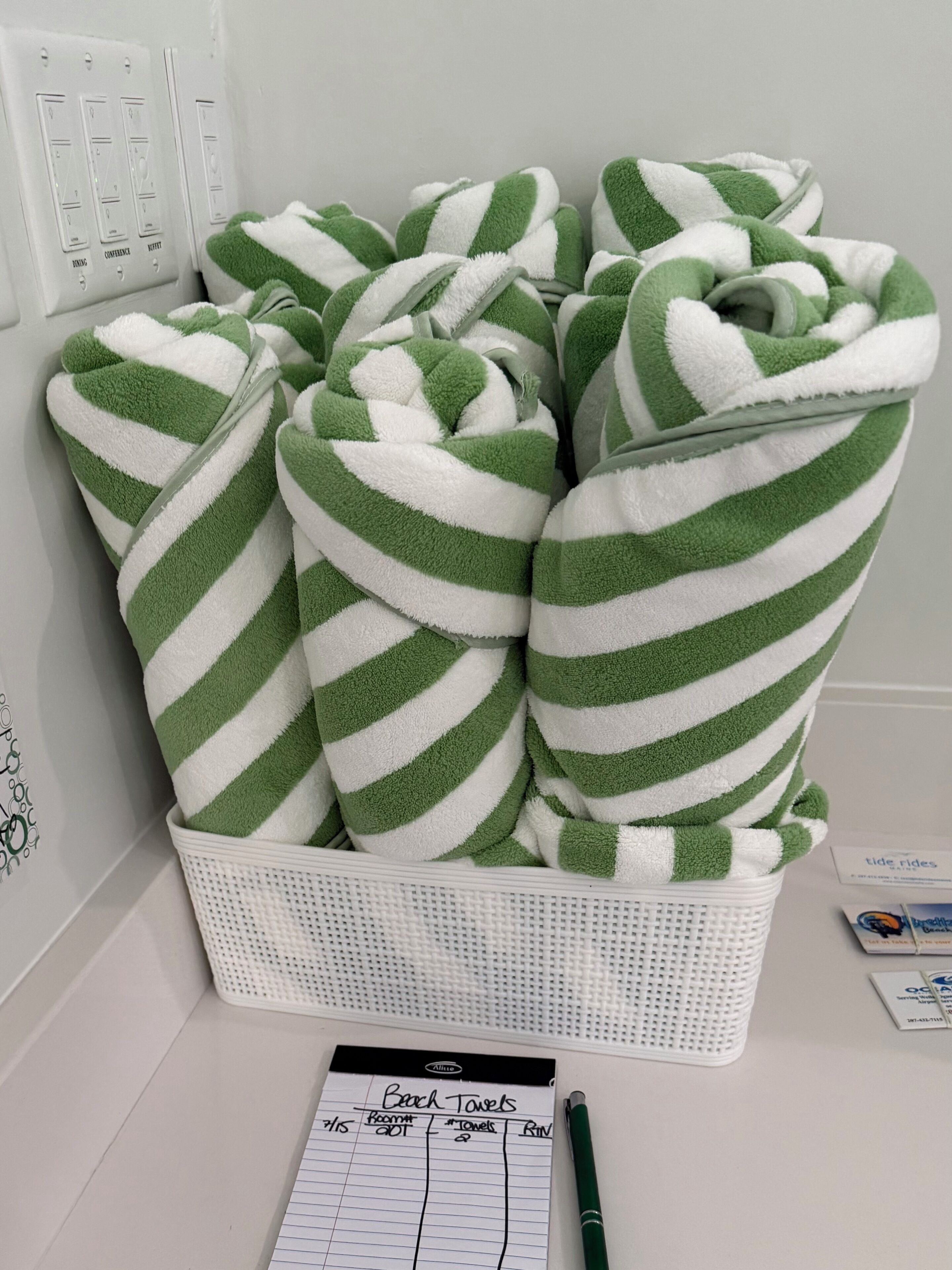 A basket of green and white striped towels, a notepad labeled 'Beach Towels,' and a pen on a white surface.