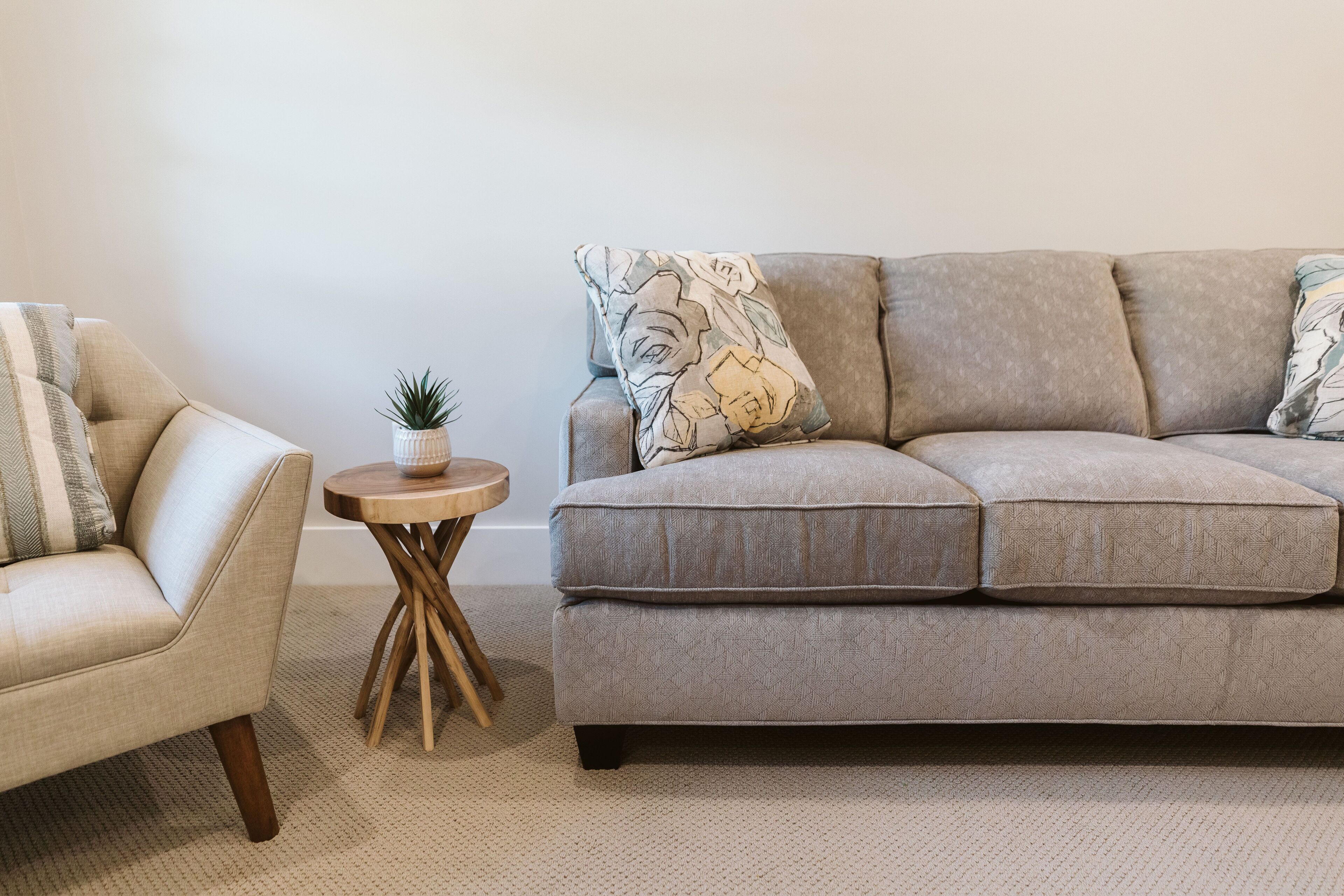 Ein Wohnzimmer mit einem beigefarbenen Sofa, einem runden Holztisch mit einer Pflanze und einem dekorativen Kissen mit Muster.