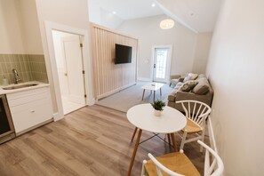 A modern kitchen with a built-in dishwasher, a dining area with a round table and white chairs, and a living area with a sofa and a TV mounted on the wall.