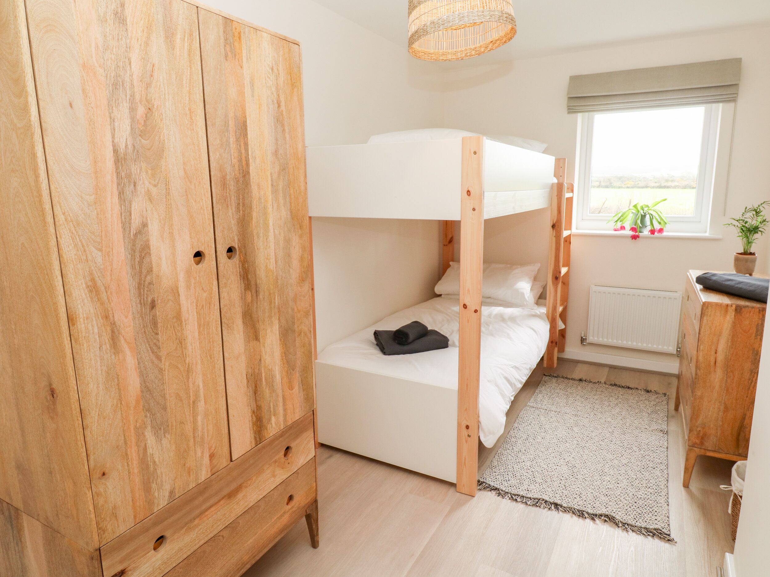 A bunk bed with a wooden wardrobe beside it, a small table, and a window with a plant.