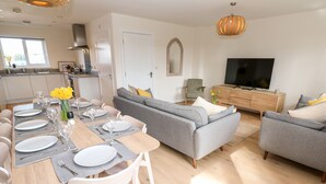 A modern living room with a grey sofa, a dining table set with plates and glasses, a TV, and a wooden cabinet. - The Bright House (Wadebridge)
