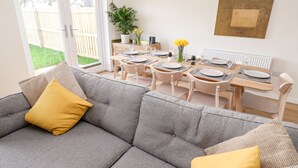 A modern living room with a grey sofa, yellow cushions, a dining area with wooden furniture, and a potted plant with yellow flowers. - The Bright House (Wadebridge)