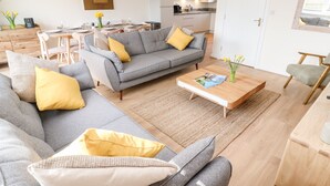 A modern living room with a grey sofa set, yellow cushions, a wooden coffee table, and a kitchen in the background. - The Bright House (Wadebridge)