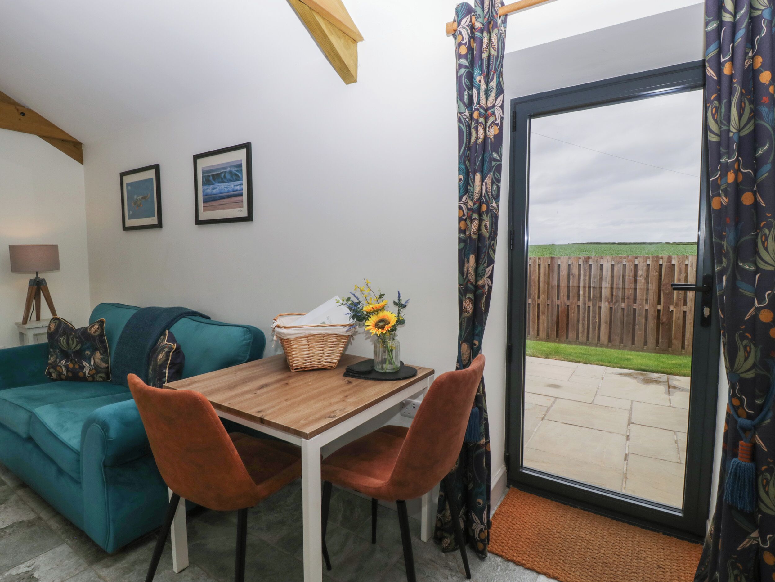 A cozy living room with a teal sofa, a wooden table, and two orange chairs. There is a wicker basket and a vase with flowers on the table. A wooden fence is visible through the glass door.