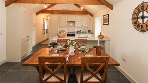 A dining area with a wooden table set for four, surrounded by wooden chairs. A kitchen is visible in the background with white cabinets and a wooden ceiling. - The Byre (Bridgend)