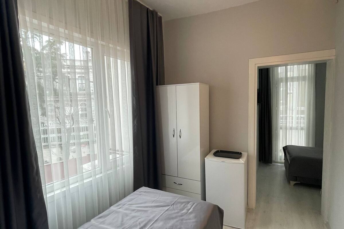 A hotel room with a bed, a wardrobe, a desk, and a chair. There is a window with curtains and a view of buildings outside.