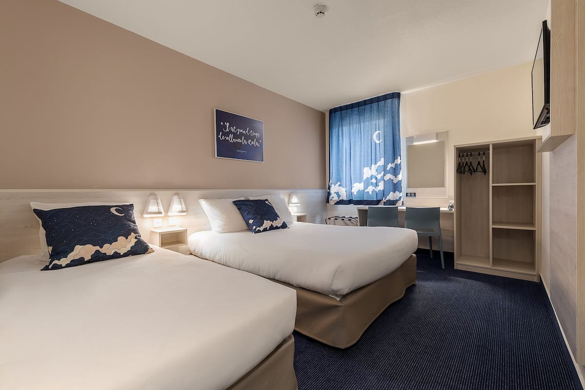 A hotel room with two beds, a desk, a chair, a wardrobe, and a wall-mounted TV.