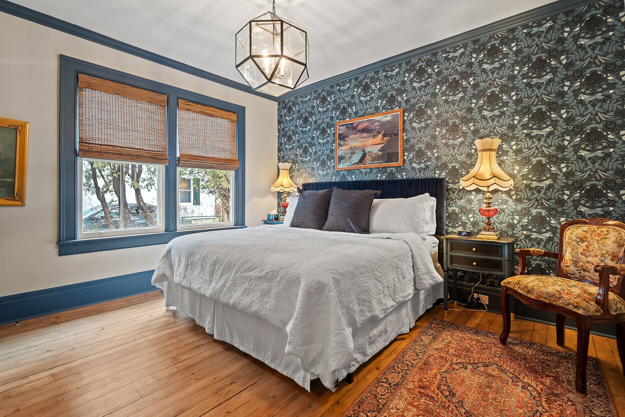 Une chambre à coucher avec un grand lit, une table de chevet, une chaise, un mur à motifs et une fenêtre avec des stores.