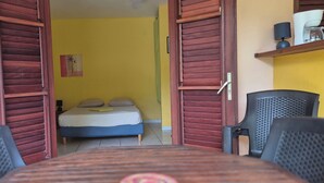 Una habitación con una cama, una mesa con sillas, un refrigerador y una estantería con una lámpara.