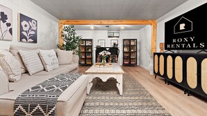 A living room with a sofa, a coffee table, and a black and white patterned rug. There is a bookshelf and a black cabinet with a logo on the wall.