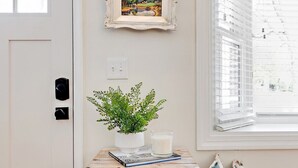 A room with a white door, a wooden dresser with drawers, a small table with a plant, a framed picture, and a window with blinds.