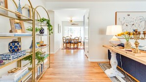 A hallway with a wooden floor leading to a dining area with a table and chairs. There are shelves with books and decorative items, and a lamp on a console table.