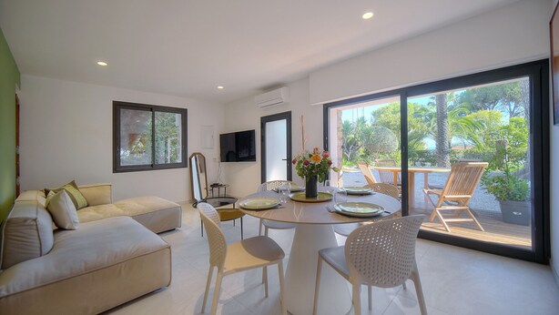 A modern living room with a sofa, dining table, and chairs. There is a TV mounted on the wall and a window with a view of the outdoors. - 2 CHAMBRES JARDIN/TERRASSE ACCES PLAGES A2B241 (Cannes)