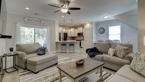 A spacious living room with a sofa, ottoman, and coffee table, a kitchen area with modern appliances, and a ceiling fan.