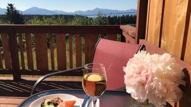 Una mesa con un plato de comida, un vaso de bebida y un florero en un balcón con baranda de madera y vistas a las montañas.