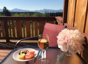 A table with a plate of food, a glass of drink, and a vase of flowers on a balcony with a wooden railing and a view of mountains.